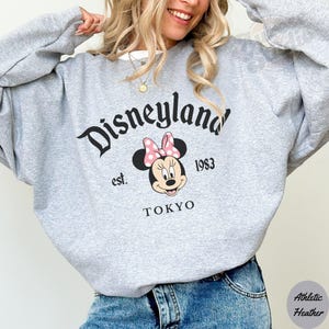 May include: A gray sweatshirt with the Disneyland logo in black script, featuring Minnie Mouse with a pink bow. The text "est. 1983 TOKYO" is below the Minnie Mouse graphic. The sweatshirt is made of athletic heather material.