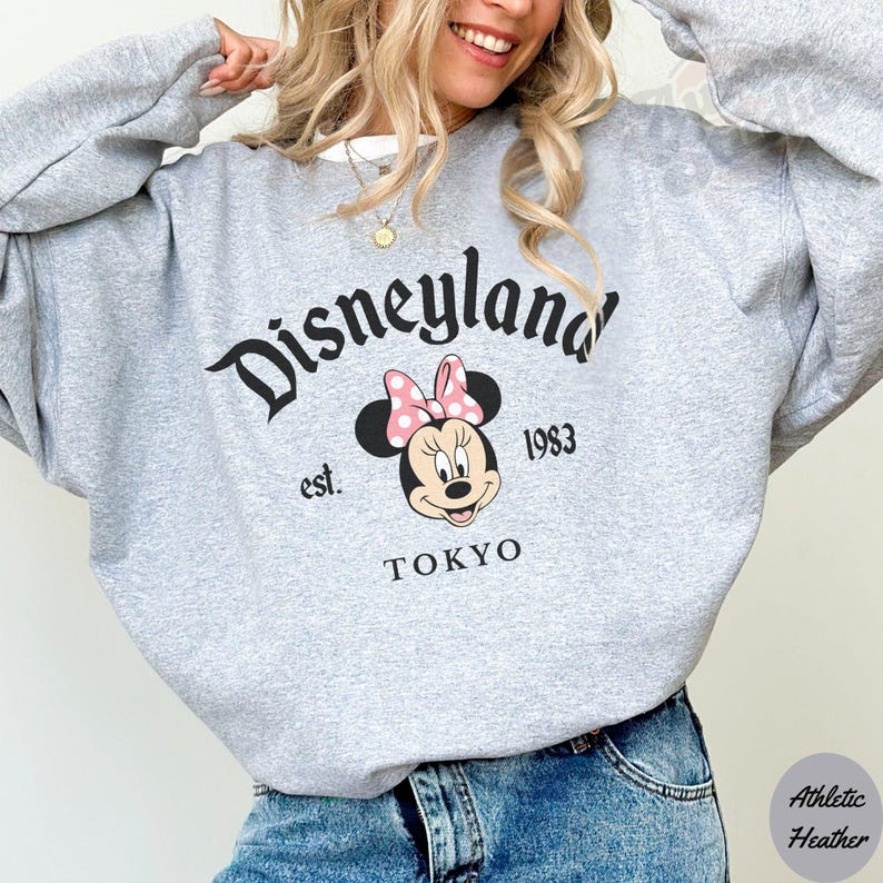 May include: A gray sweatshirt with the Disneyland logo in black script, featuring Minnie Mouse with a pink bow. The text "est. 1983 TOKYO" is below the Minnie Mouse graphic. The sweatshirt is made of athletic heather material.