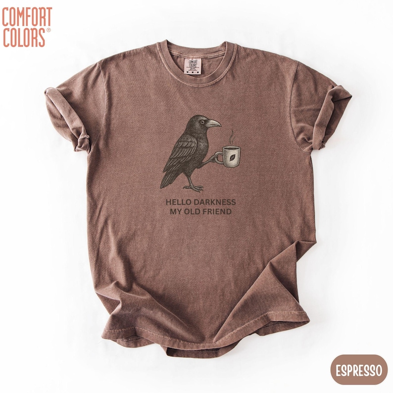 May include: A brown short-sleeve t-shirt featuring a graphic of a black crow holding a steaming mug. Below the crow, the text reads, "HELLO DARKNESS MY OLD FRIEND." The shirt has a crew neckline and a relaxed fit, with the brand name Comfort Colors visible.