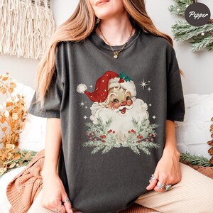 May include: Dark gray t-shirt featuring a vintage Santa Claus graphic. Santa is depicted with a red hat, white beard, and a smiling face, surrounded by greenery and berries. The shirt is a comfortable, casual style.