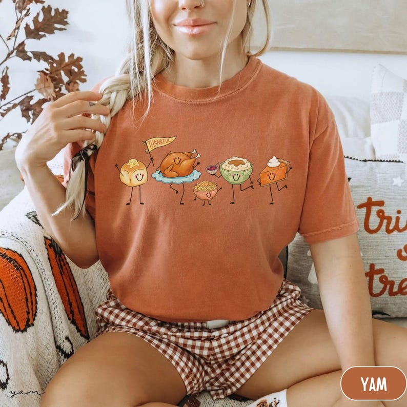 May include: A rust-colored short-sleeve t-shirt featuring a graphic print of cartoon Thanksgiving food items with smiling faces, including a roasted turkey holding a flag that says Thankful, a dinner roll, mashed potatoes, and a slice of pumpkin pie.