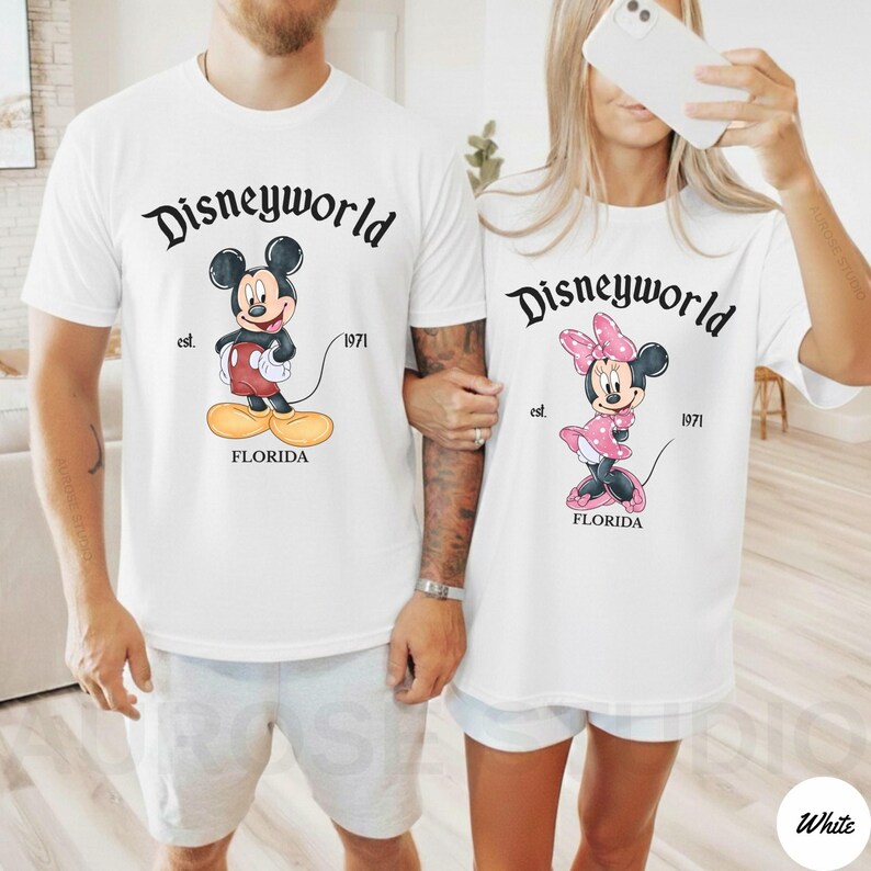 May include: White t-shirts featuring Mickey and Minnie Mouse, with "Disneyworld" in a script font above the characters. The shirts also include "est. 1971" and "FLORIDA" in a serif font. The shirts are worn by two people.