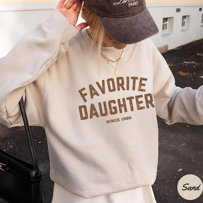 K&ouml;nnte beinhalten: Ein sandfarbener Pullover mit dem Text "FAVORITE DAUGHTER SINCE 1990" in braunen Blockbuchstaben. Der Pullover ist ein l&auml;ssiger Stil, und das Bild zeigt auch eine dunkelbraune Kappe.