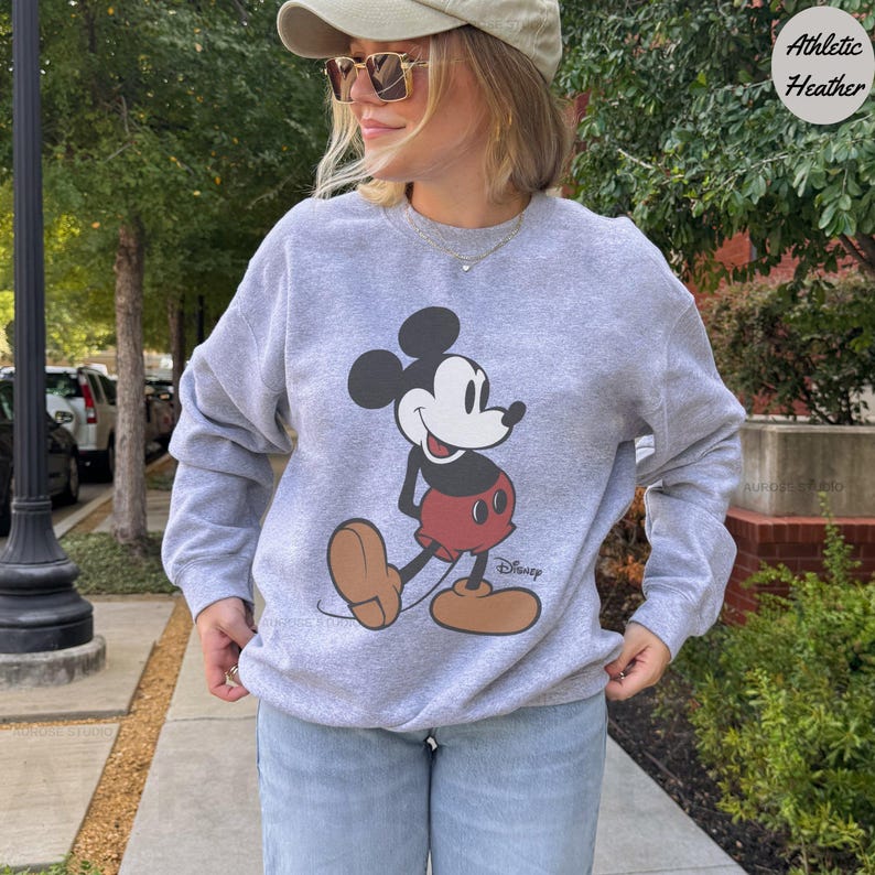 Puede incluir: Sudadera gris jaspeado con un gran gr&aacute;fico de Mickey Mouse en su pose cl&aacute;sica. El logotipo de Disney est&aacute; impreso debajo del personaje. La sudadera es de manga larga y tiene cuello redondo.