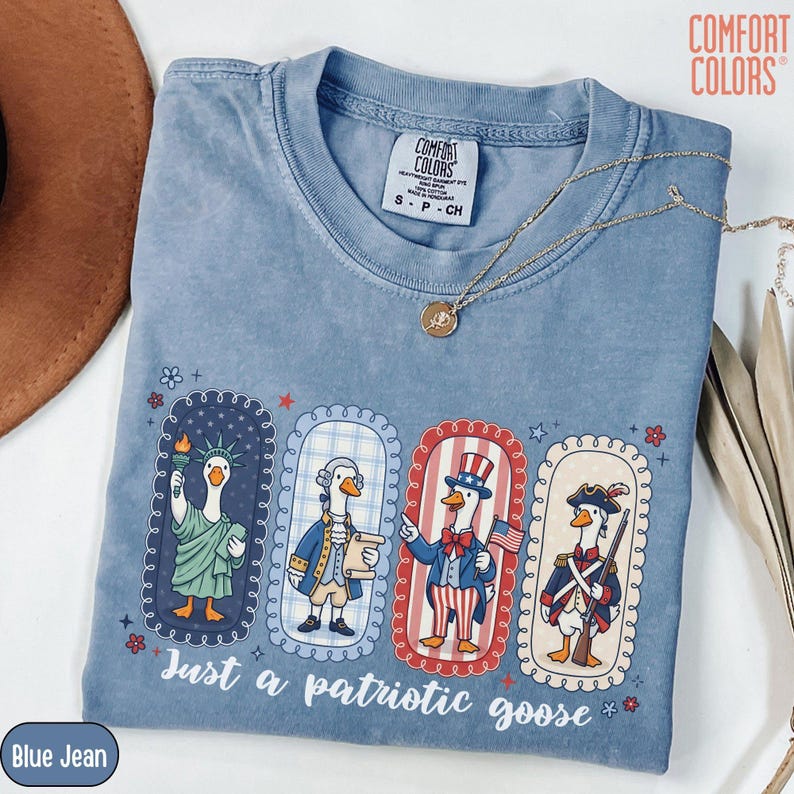 May include: Blue jean Comfort Colors t-shirt featuring a graphic of patriotic geese. The geese are dressed as the Statue of Liberty, a Founding Father, Uncle Sam, and a Revolutionary War soldier. The text reads "Just a patriotic goose."