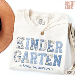 May include: An off-white t-shirt with the word KINDERGARTEN printed in blue patchwork patterns featuring floral, gingham, and polka dot designs. Below the text, the name Mrs. Anderson is printed in blue script, flanked by small daisy icons.