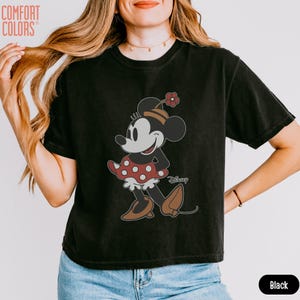 May include: Black t-shirt featuring a cartoon Minnie Mouse design. Minnie Mouse is wearing a red and white polka dot skirt, brown shoes, and a hat with a flower. The word "Disney" is printed on the shirt.