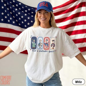 May include: White t-shirt with a patriotic goose design. The graphic features four geese dressed in patriotic attire, with the text "Just a patriotic goose". A blue "USA" cap and an American flag are in the background.