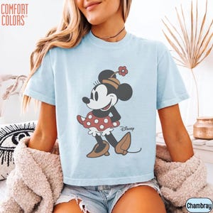 May include: Light blue t-shirt featuring a Minnie Mouse graphic. The graphic shows Minnie Mouse in a red polka dot dress and a hat with a flower. The shirt has the "Comfort Colors" logo on the upper left and the Disney logo on the lower right.