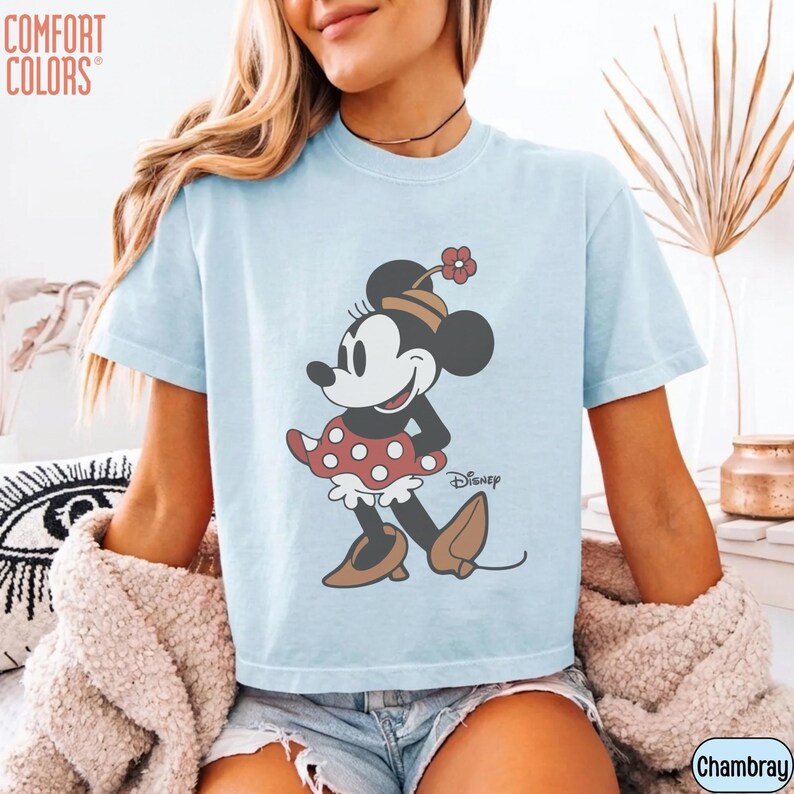 May include: Light blue t-shirt featuring a Minnie Mouse graphic. The graphic shows Minnie Mouse in a red polka dot dress and a hat with a flower. The shirt has the "Comfort Colors" logo on the upper left and the Disney logo on the lower right.