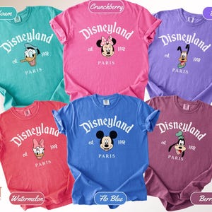 May include: A collection of six colorful t-shirts featuring the Disneyland logo and various Disney characters. Each shirt displays "Disneyland Paris" and the year "1992". The shirts come in seafoam, crunchberry, violet, watermelon, flo blue, and berry.