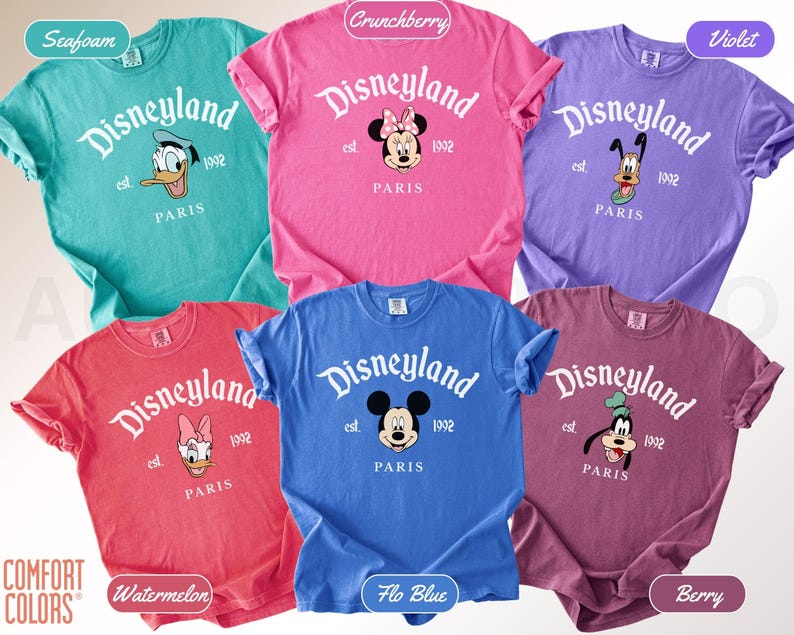 May include: A collection of six colorful t-shirts featuring the Disneyland logo and various Disney characters. Each shirt displays "Disneyland Paris" and the year "1992". The shirts come in seafoam, crunchberry, violet, watermelon, flo blue, and berry.