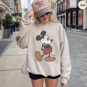 Puede incluir: Sudadera beige con un gran dise&ntilde;o de Mickey Mouse en negro, rojo y marr&oacute;n. La sudadera tiene cuello redondo y mangas largas. El logotipo de Disney est&aacute; impreso debajo del personaje. Tambi&eacute;n se ve una gorra.
