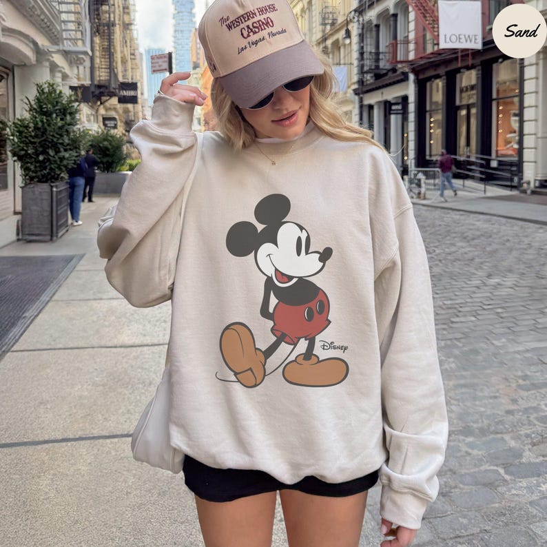 Puede incluir: Sudadera beige con un gran dise&ntilde;o de Mickey Mouse en negro, rojo y marr&oacute;n. La sudadera tiene cuello redondo y mangas largas. El logotipo de Disney est&aacute; impreso debajo del personaje. Tambi&eacute;n se ve una gorra.