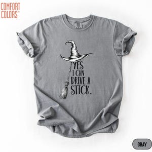 Puede incluir: Una camiseta gris de manga corta con un gr&aacute;fico negro de un sombrero de bruja sobre una escoba. El texto dice, YES I CAN DRIVE A STICK. La prenda tiene un cuello redondo y una etiqueta de la marca Comfort Colors impresa en el interior.