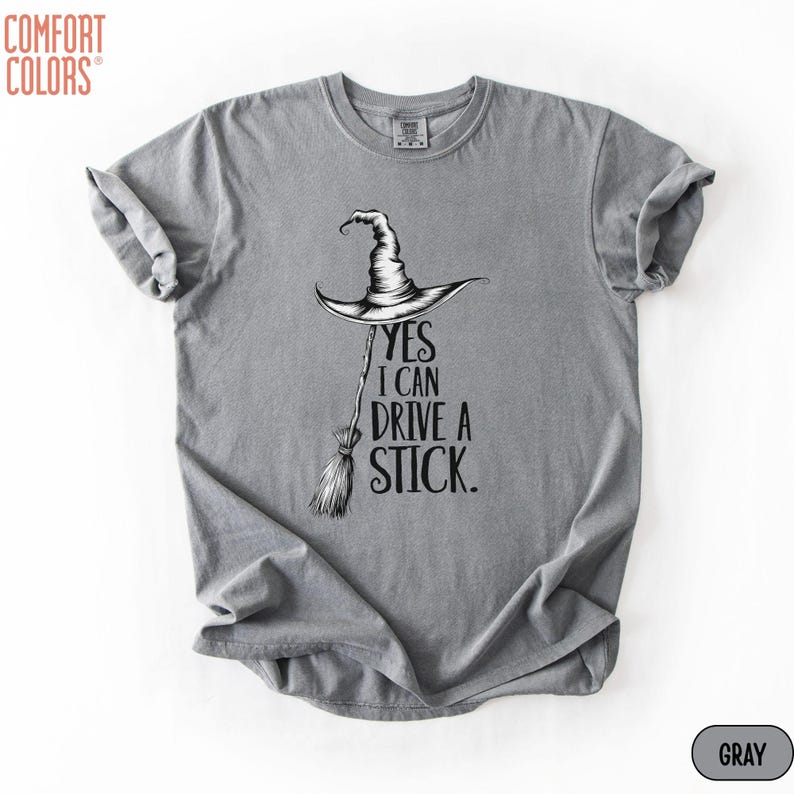 Puede incluir: Una camiseta gris de manga corta con un gr&aacute;fico negro de un sombrero de bruja sobre una escoba. El texto dice, YES I CAN DRIVE A STICK. La prenda tiene un cuello redondo y una etiqueta de la marca Comfort Colors impresa en el interior.