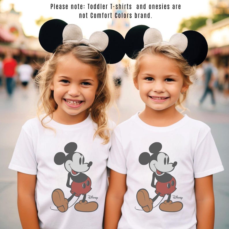 May include: Two young girls wearing white t-shirts featuring a Mickey Mouse graphic. They are also wearing black and tan mouse ear headbands. The image includes the text: "Please note: Toddler T-shirts and onesies are not Comfort Colors brand."