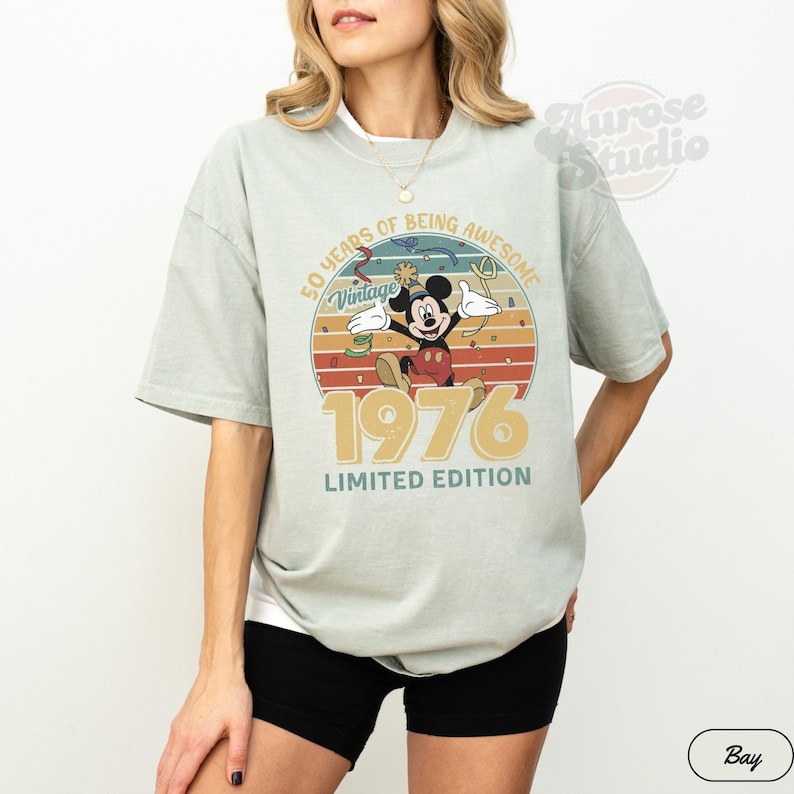 Puede incluir: Camiseta verde claro con un dise&ntilde;o vintage de Mickey Mouse. El gr&aacute;fico incluye el texto "50 YEARS OF BEING AWESOME", "Vintage", "1976" y "LIMITED EDITION" en un estilo retro. La camiseta es de corte holgado.