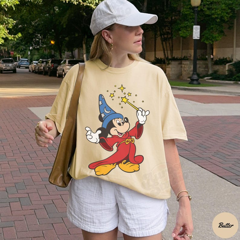 May include: A light yellow t-shirt featuring a graphic of Mickey Mouse dressed as a wizard, holding a wand with stars. The shirt is worn with white shorts and a white baseball cap. The shirt has short sleeves.