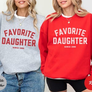 K&ouml;nnte beinhalten: Zwei Sweatshirts, eines in Athletic Heather Grau und eines in Rot, beide mit dem Text "FAVORITE DAUGHTER SINCE 1990" in verblichenen roten und wei&szlig;en Buchstaben. Das graue Sweatshirt wird mit Jeans getragen, das rote mit schwarzen Shorts.