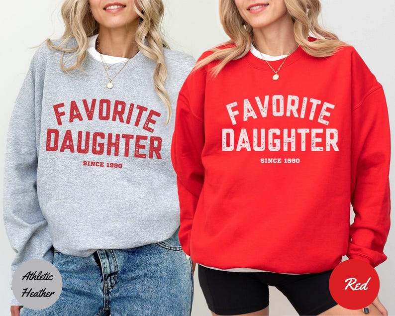 K&ouml;nnte beinhalten: Zwei Sweatshirts, eines in Athletic Heather Grau und eines in Rot, beide mit dem Text "FAVORITE DAUGHTER SINCE 1990" in verblichenen roten und wei&szlig;en Buchstaben. Das graue Sweatshirt wird mit Jeans getragen, das rote mit schwarzen Shorts.
