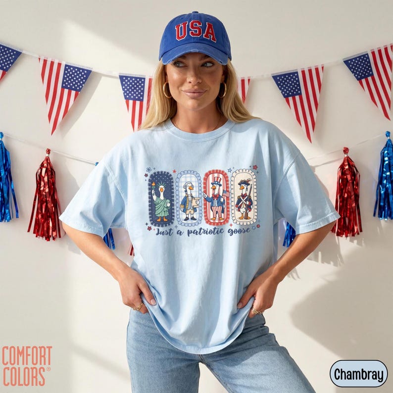 May include: Pale blue t-shirt featuring a patriotic goose design and the text "Just a patriotic goose." The design shows four geese in patriotic outfits. A blue baseball cap with "USA" is worn.