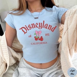 Puede incluir: Top corto azul claro con la palabra "Disneyland" en escritura roja, junto con "est. 1955" y "CALIFORNIA". El dise&ntilde;o incluye un gr&aacute;fico de cereza con un lazo rosa. La camiseta tiene mangas cortas.