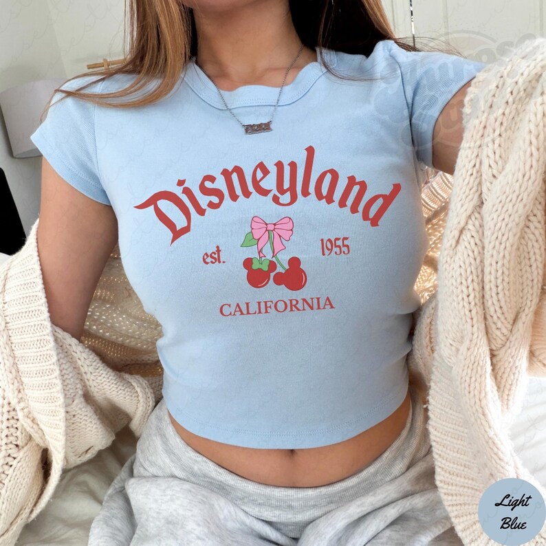 Puede incluir: Top corto azul claro con la palabra "Disneyland" en escritura roja, junto con "est. 1955" y "CALIFORNIA". El dise&ntilde;o incluye un gr&aacute;fico de cereza con un lazo rosa. La camiseta tiene mangas cortas.