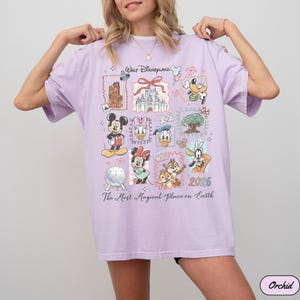K&ouml;nnte beinhalten: Orchideenfarbenes T-Shirt mit einem Disney-Motiv. Das Design zeigt verschiedene Disney-Charaktere und Wahrzeichen, wie Micky Maus, Minnie Maus und das Schloss von Cinderella. Der Text "Walt Disneyland" und "The Most Magical Place on Earth" sind ebenfalls zu sehen.