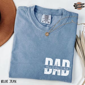 May include: A blue jean t-shirt with a white graphic that says "DAD" with the names "Oliver, Liam, Noah" underneath. The shirt has a distressed look and is made of a soft, comfortable fabric.