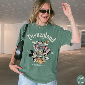 May include: Light green t-shirt with the text "Disneyland" and a graphic of Disney characters. The shirt includes the text "est. 1955" and "CALIFORNIA". The shirt is a short-sleeved crew neck.