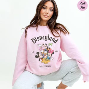 K&ouml;nnte beinhalten: Hellrosa Disneyland-Sweatshirt mit Mickey Mouse, Minnie Mouse, Donald Duck, Daisy Duck, Goofy und Pluto. Das Sweatshirt hat den Text "Disneyland", "est. 1955" und "CALIFORNIA".