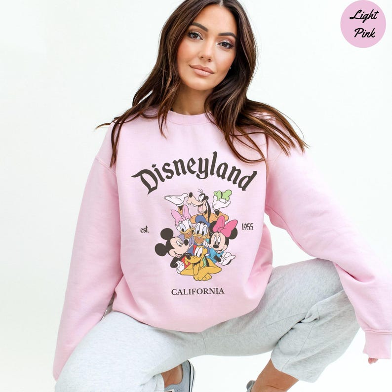 K&ouml;nnte beinhalten: Hellrosa Disneyland-Sweatshirt mit Mickey Mouse, Minnie Mouse, Donald Duck, Daisy Duck, Goofy und Pluto. Das Sweatshirt hat den Text "Disneyland", "est. 1955" und "CALIFORNIA".