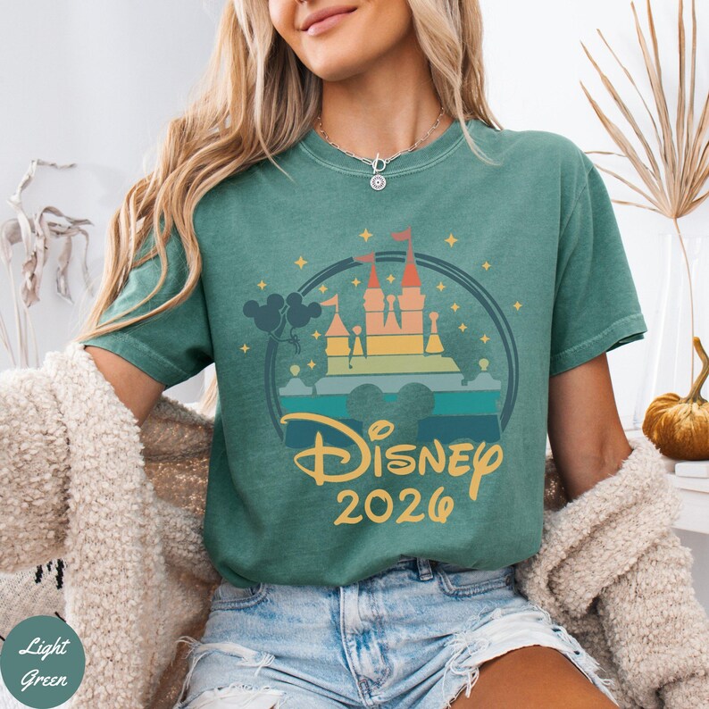 May include: Light green t-shirt with a colorful graphic of a castle, Mickey Mouse ears, and the text "Disney 2026". The design is within a circular frame. The shirt is a solid color.