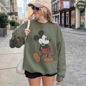 Puede incluir: Sudadera verde militar con un gr&aacute;fico de Mickey Mouse. El dise&ntilde;o incluye la cara de Mickey, pantalones cortos rojos y zapatos color canela. El logotipo de Disney est&aacute; impreso debajo. La sudadera es de color s&oacute;lido y tiene mangas largas.
