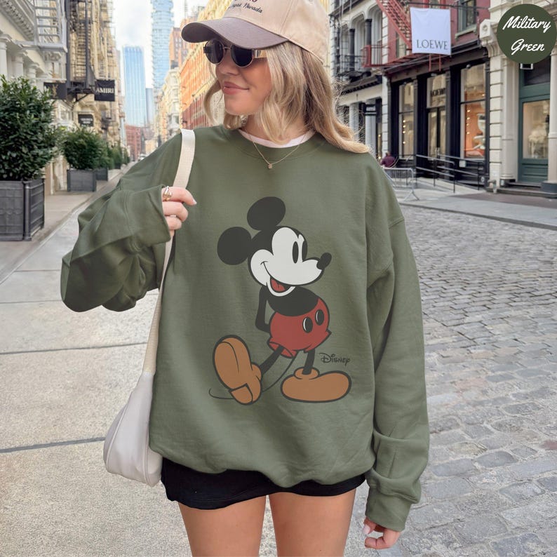 Puede incluir: Sudadera verde militar con un gr&aacute;fico de Mickey Mouse. El dise&ntilde;o incluye la cara de Mickey, pantalones cortos rojos y zapatos color canela. El logotipo de Disney est&aacute; impreso debajo. La sudadera es de color s&oacute;lido y tiene mangas largas.