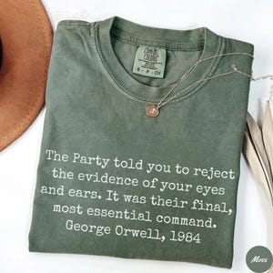 Könnte beinhalten: Olivgrünes T-Shirt mit dem Zitat "The Party told you to reject the evidence of your eyes and ears. It was their final, most essential command. George Orwell, 1984" in Weiß. Eine goldene Halskette mit Anhänger liegt auf dem Shirt.