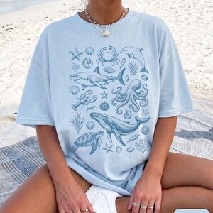 Pode incluir: Uma t-shirt azul claro com uma impress&atilde;o gr&aacute;fica azul escura de v&aacute;rias criaturas marinhas, incluindo um tubar&atilde;o, baleia, polvo, tartaruga marinha, caranguejo, golfinho, lagosta e v&aacute;rias conchas e corais, com a palavra CHAMBRAY no canto.