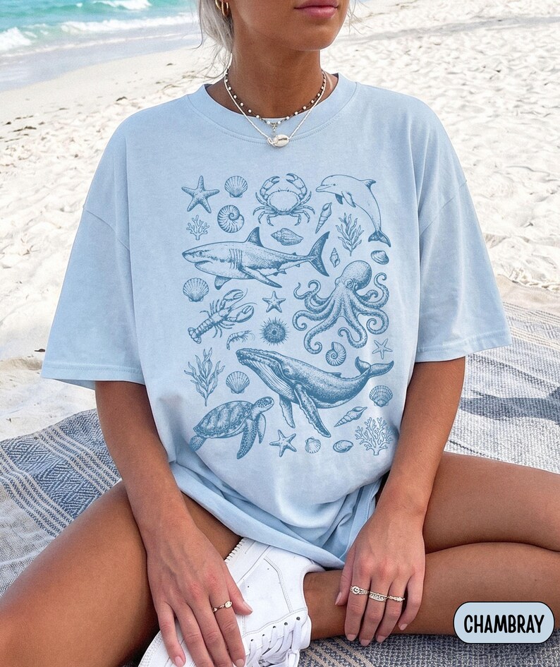 Pode incluir: Uma t-shirt azul claro com uma impress&atilde;o gr&aacute;fica azul escura de v&aacute;rias criaturas marinhas, incluindo um tubar&atilde;o, baleia, polvo, tartaruga marinha, caranguejo, golfinho, lagosta e v&aacute;rias conchas e corais, com a palavra CHAMBRAY no canto.