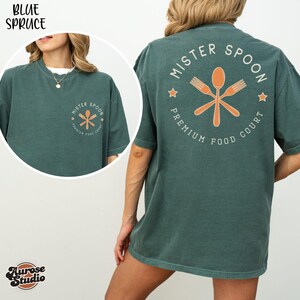 May include: A teal-colored t-shirt with the text "MISTER SPOON PREMIUM FOOD COURT" in a circular design on the back. The design includes a crossed spoon and forks in orange. The front of the shirt has a smaller version of the logo.