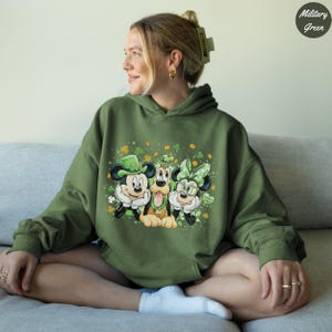 May include: Military green hoodie featuring Mickey Mouse, Pluto, and Minnie Mouse in St. Patrick's Day attire. Mickey wears a green hat, Minnie a green bow, and Pluto has a green shamrock. The design includes shamrocks and the words "Lucky Me".