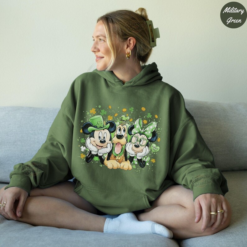 May include: Military green hoodie featuring Mickey Mouse, Pluto, and Minnie Mouse in St. Patrick's Day attire. Mickey wears a green hat, Minnie a green bow, and Pluto has a green shamrock. The design includes shamrocks and the words "Lucky Me".