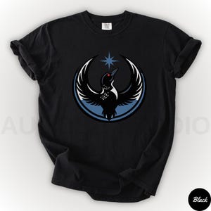 K&ouml;nnte beinhalten: Schwarzes T-Shirt mit einer Grafik eines Vogels mit ausgebreiteten Fl&uuml;geln, einem Stern und einem blauen Kreis. Der Vogel hat ein rotes Auge und ist vor einem wei&szlig;en und blauen Hintergrund platziert. Das T-Shirt ist ein l&auml;ssiger Stil.