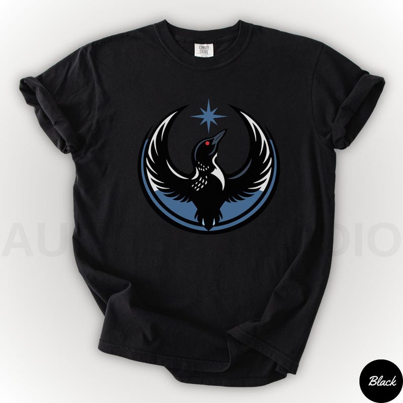 K&ouml;nnte beinhalten: Schwarzes T-Shirt mit einer Grafik eines Vogels mit ausgebreiteten Fl&uuml;geln, einem Stern und einem blauen Kreis. Der Vogel hat ein rotes Auge und ist vor einem wei&szlig;en und blauen Hintergrund platziert. Das T-Shirt ist ein l&auml;ssiger Stil.
