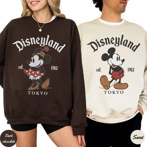 Puede incluir: Dos sudaderas, una de chocolate oscuro y otra color arena, con el logotipo de Disneyland con Mickey y Minnie Mouse. Las sudaderas tienen el texto "Disneyland est. 1983 TOKYO". La sudadera de Minnie Mouse es de chocolate oscuro, y la sudadera de Mickey Mouse es color arena.