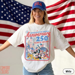 May include: White t-shirt with a colorful graphic celebrating the United States of America's 250th anniversary. The design includes the text "Anniversary 250 Years of Freedom" and features cartoon characters. A blue baseball cap with "USA" is also visible.
