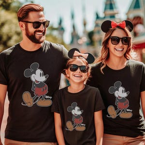 May include: Three people wearing black t-shirts featuring a Mickey Mouse graphic. The cartoon character is depicted in a classic pose with red shorts, brown shoes, and white gloves. The family is smiling and wearing sunglasses.