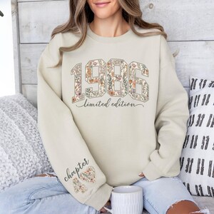 May include: Beige sweatshirt with floral-filled "1986" graphic and "limited edition" text. The left sleeve features "chapter 40" and a floral heart. The sweatshirt is a neutral color, suitable for various occasions.