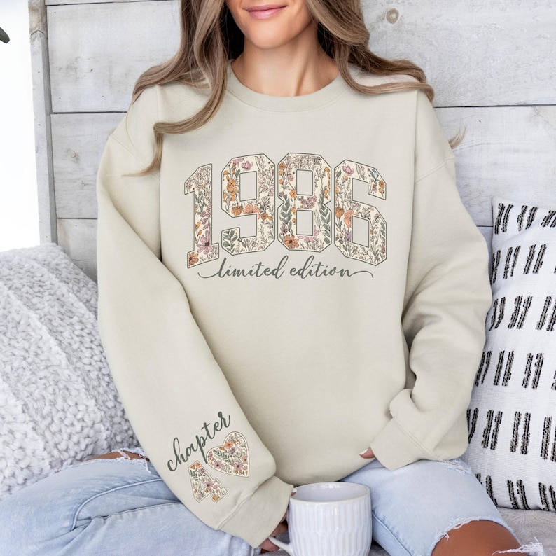 May include: Beige sweatshirt with floral-filled "1986" graphic and "limited edition" text. The left sleeve features "chapter 40" and a floral heart. The sweatshirt is a neutral color, suitable for various occasions.