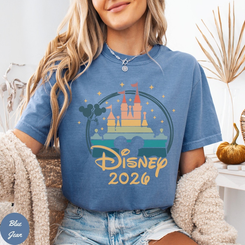 May include: Blue jean-colored t-shirt featuring a colorful castle graphic with the text "Disney 2026". The graphic includes a castle, balloons, and a circular design. The shirt is a comfortable, casual style.